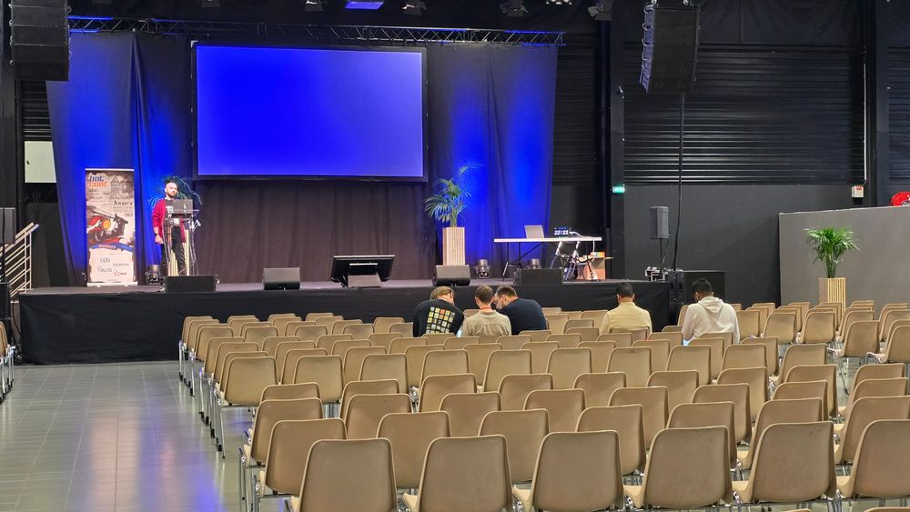 View of the Botconf 2025 conference with a large blue screen at the top. Mostly empty seats for now, 5 people can be seen.
