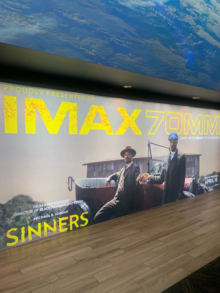 A poster for Sinners the Movie, directed by Ryan Coogler and starring Michael B. Jordan. Two of the movies characters stand in front of an old timey red car. The text above says "Proudly Presented in IMAX 70MM"