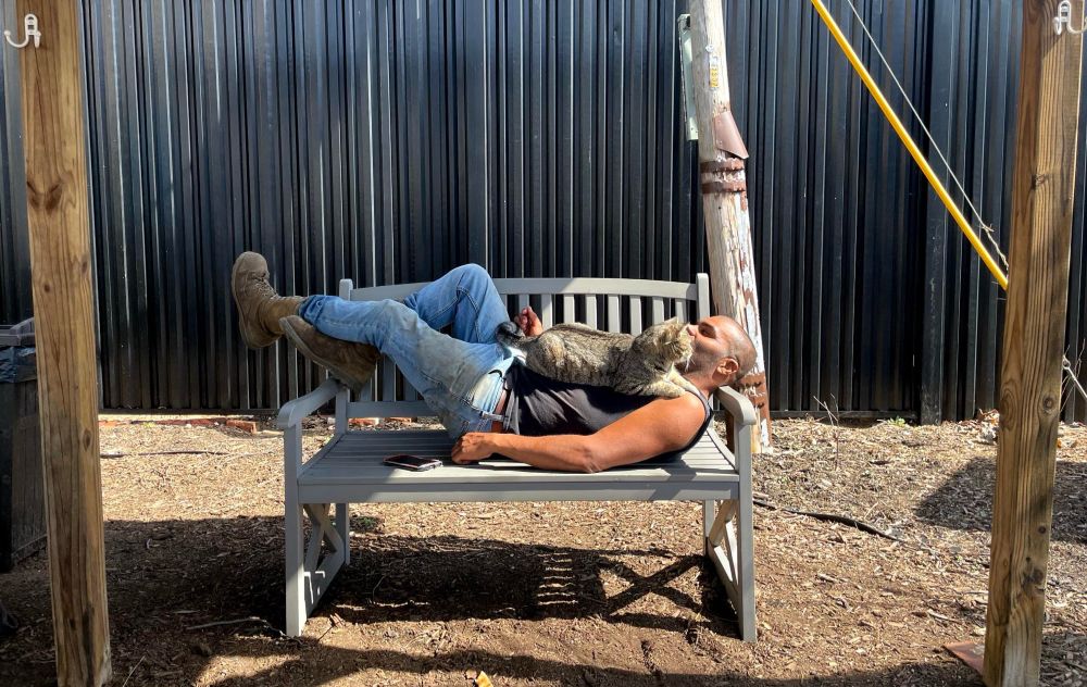 My partner (a devastatingly handsome Black man in work boots and a shaved head) napping on a bench with a cat sitting on his chest 