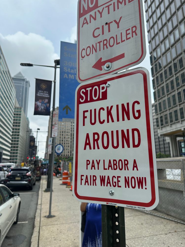A white sign reads “stop fucking around, pay labor a fair wage now!”