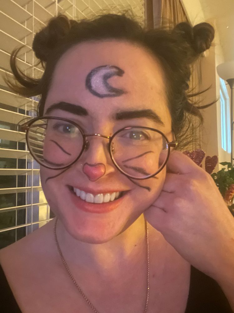 A selfie of a brunette woman (me), wearing glasses, with her hair in space buns that were supposed to kinda be cat ears but are sorta falling over. 

Nose painted pink & outlined, sort of crooked whiskers, and a very poorly drawn moon on my forehead as a nod to Luna from Sailor Moon. Eyebrows over-lined for emphasis. 

I normally wear contacts and since I’m pretty dang blind, drawing this stuff on while my glasses were off was not easy 🫣😂

I never take pics of myself anymore, feels awkward to for some reason?