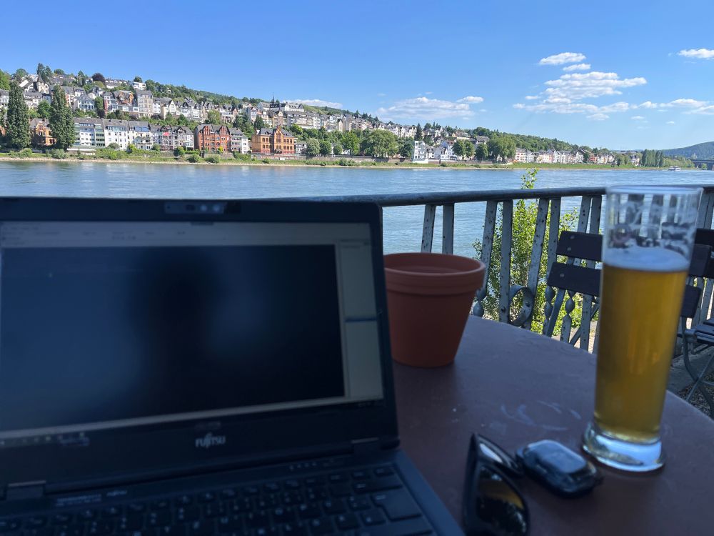 Ein Flussufer. Auf der gegenüberliegenden Seite ist Sonne satt. Auf einem Gartentisch eines biergartens steht ein laptop und ein alkoholfreies weizenbier. 