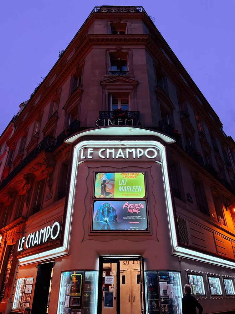 The facade of Le Champo cinema, colloquially known as Espace Jacque Tati, lit up in white neon against a twilight sky glowing blue and purple. 