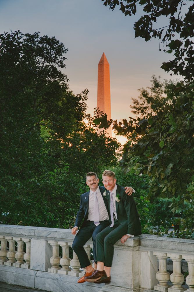 PT and G sitting on a balcony in their wedding suits with the Washington monument rising in the sunset behind them. 