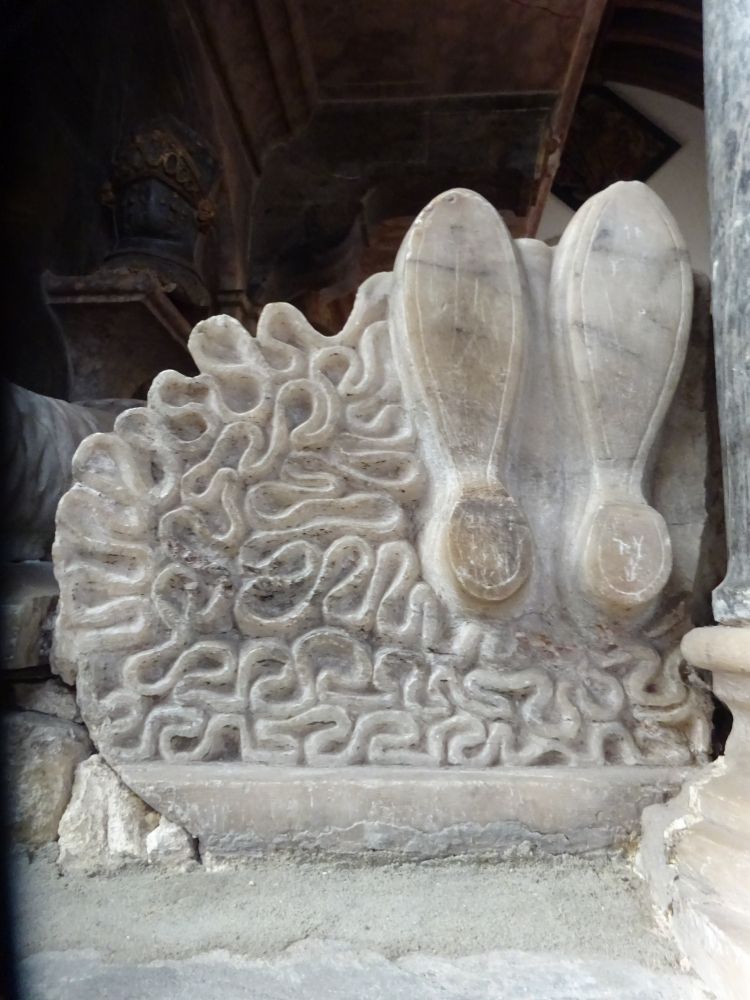 The side of an woman's effigy tomb showing the bottom of her dress and her feet (she lays on her back). The dress is depicted as many wiggled folds of fabric, and we can see the soles of her small shoes. 