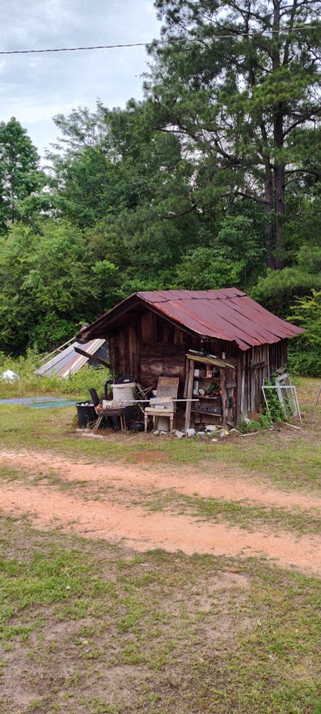 Picture of a worn down wooden utility shed with a rusted roof. There is a dirt road in front of it and a grove of trees behind it. 