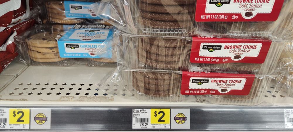A shelf at a local Dollar General with Clover Valley store brand soft baked cookies listed for $2 a package, 8 cookies to a package. On the left sits chocolate chip cookies, on the right sits 'brownie' cookies. 