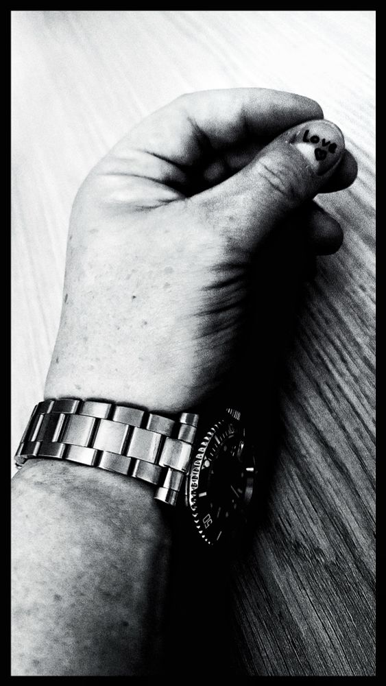 Schwarzweißaufnahme, hart kontrastiert. Zu sehen ist ein Unterarm mit einer schweren Metallarmbanduhr. Die Haut wirkt körnig, fast filmisch – feine Flecken, leichte Falten, klare Textur. Die Hand ist locker zur Faust geschlossen und liegt auf einer glatten Holzoberfläche.

Der Blickfang sitzt auf dem Daumennagel: Darauf steht handgeschrieben „Love“ mit einem kleinen Herz daneben. Durch den starken Kontrast wirkt der Schriftzug fast wie ein Tattoo.

Die Uhr dominiert das untere Bilddrittel – massives Edelstahlband, großes rundes Gehäuse, Zackenlünette. Das Motiv erinnert an analoge Street-Fotografie: roh, direkt, ein bisschen melancholisch. Die schwarze Rahmenkante hält das Ganze wie in einem klassischen Fotoabzug zusammen.