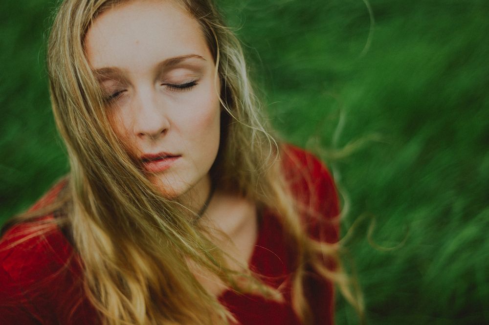 Portrait einer jungen, blonden Frau mit langen, wehenden Haaren und geschlossenen Augen und ganz leicht geöffnetem Mund, von oben gesehen, die ein rotes Oberteil trägt und vor einem grünen, unscharfen Hintergrund festgehalten ist.
