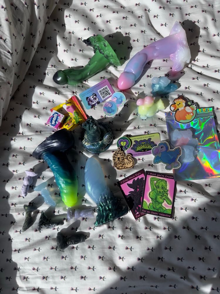 Scattered package contents over bedspread, including several dongles, teenies and squish, stickers, a wooden charm, candy, and business cards from shops.