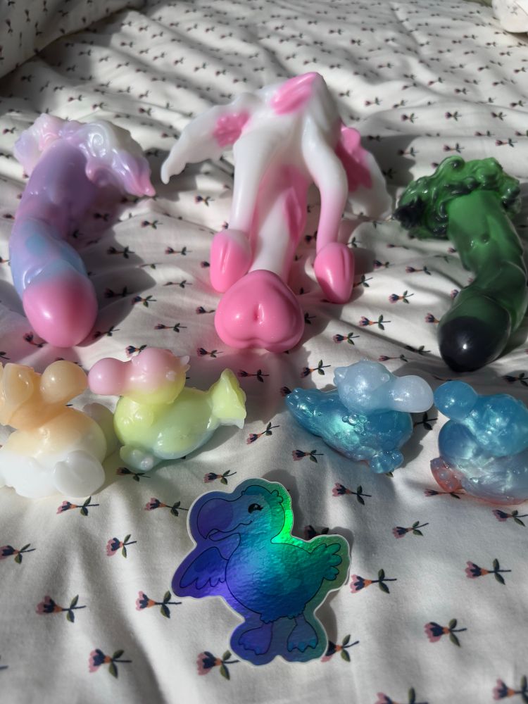 A collection of three dongles, four dodo squish, and a holographic dodo sticker on floral bedspread. The two dodos of similar pours are beak to beak.