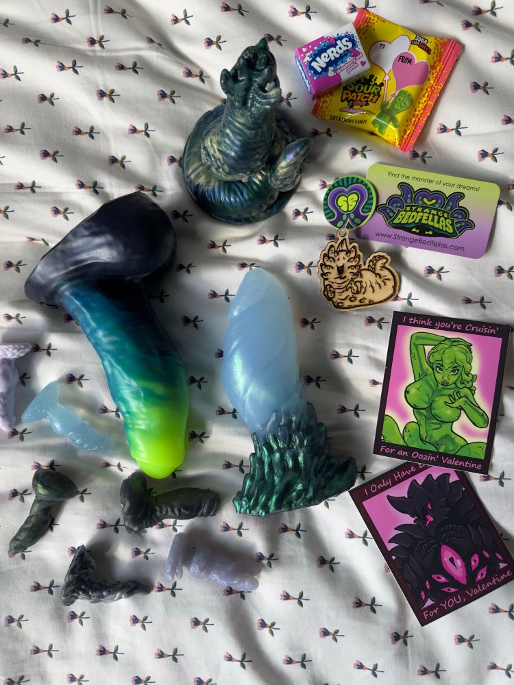 Scattered package contents from SBF over bedspread, including several dongles, teenies, stickers, a wooden charm, candy, and business card