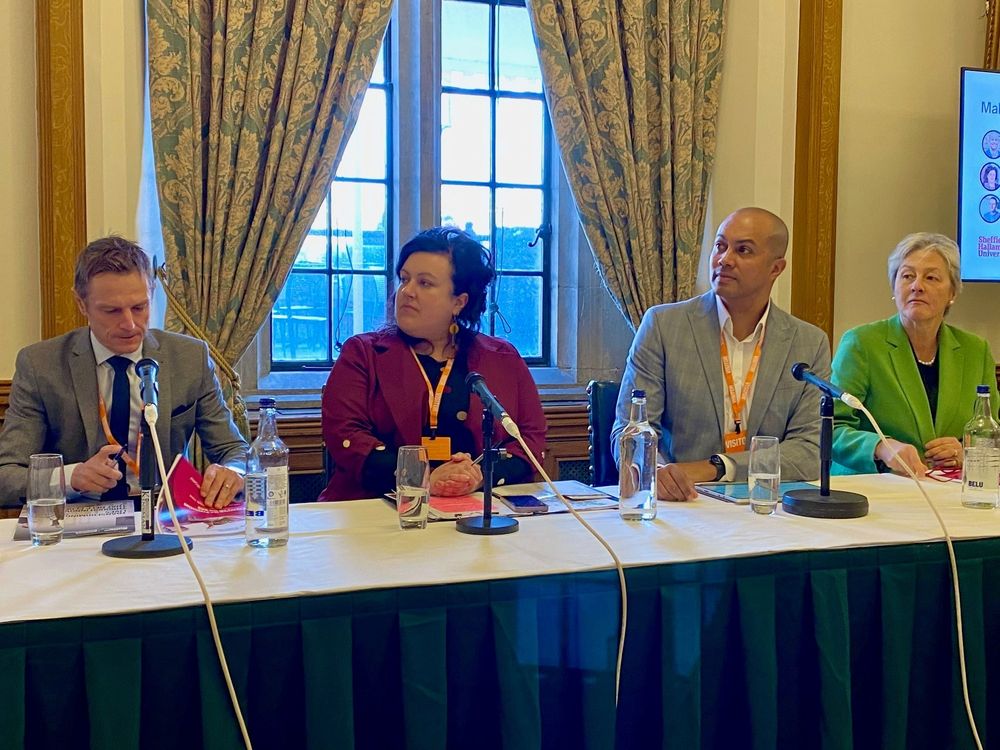 Four people sat in a room at a desk with microphones in front of them and a large window behind them 