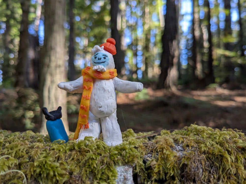 A miniature Otto the Abominable Snow Dancer hikes on a mossy log, with trees and trees for days in the background. He keeps hydrated with a big bottle of water