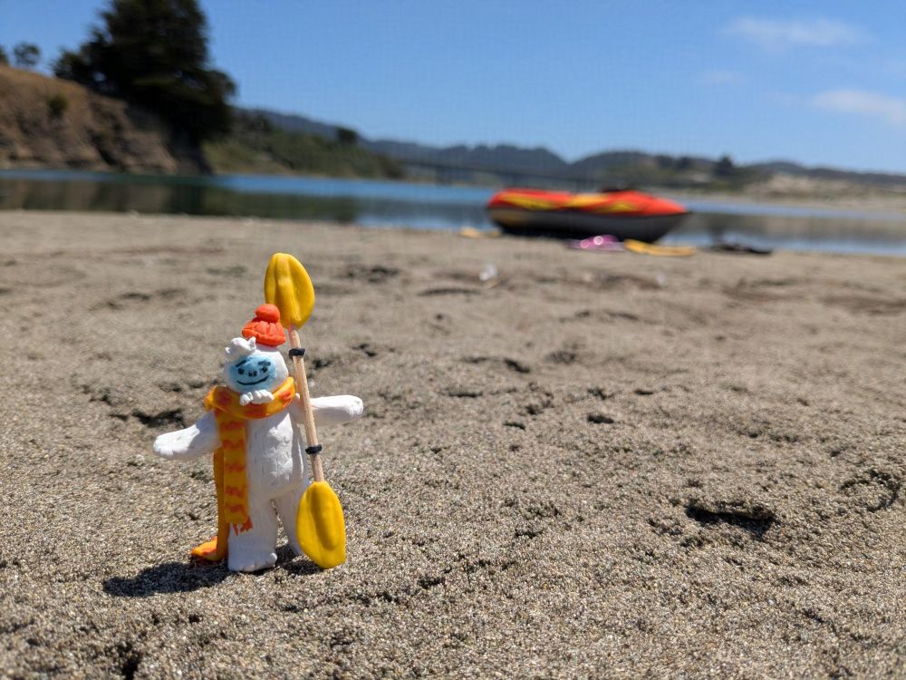 Miniature Otto the Abominable Snow Dancer holds a kayak paddle on the beach. In the background is an orange kayak and a waterway.