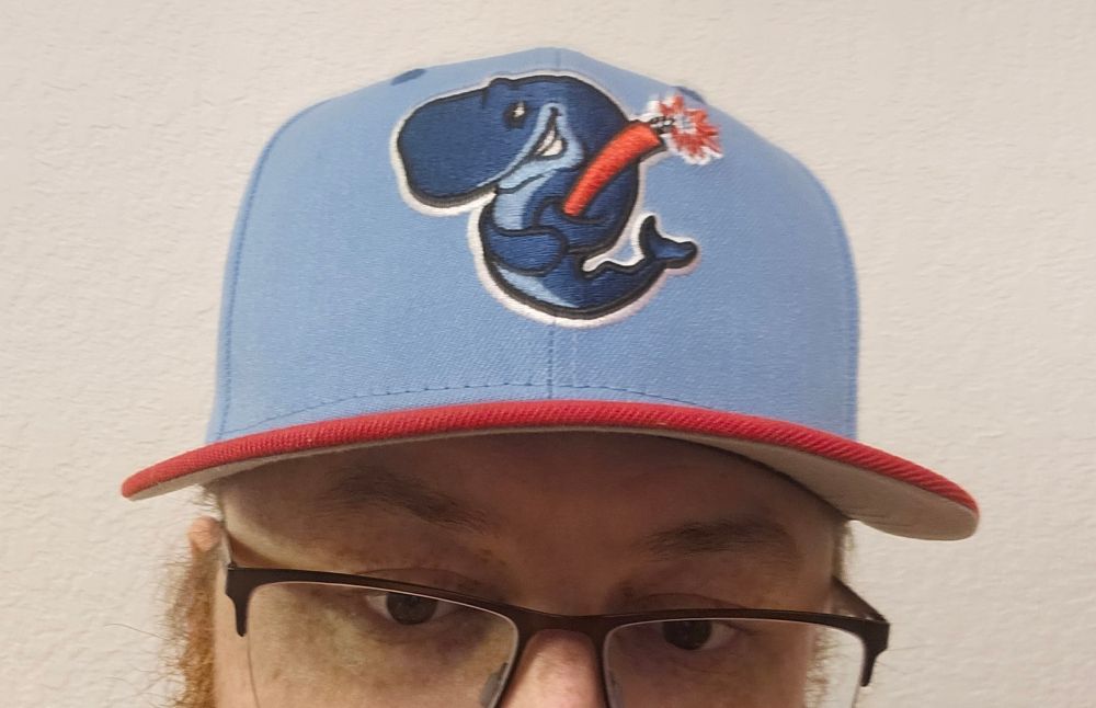 A pale blue baseball hat with a red brim. The logo on the hat is a smirking whale holding a lit stick of dynamite like a baseball bat.