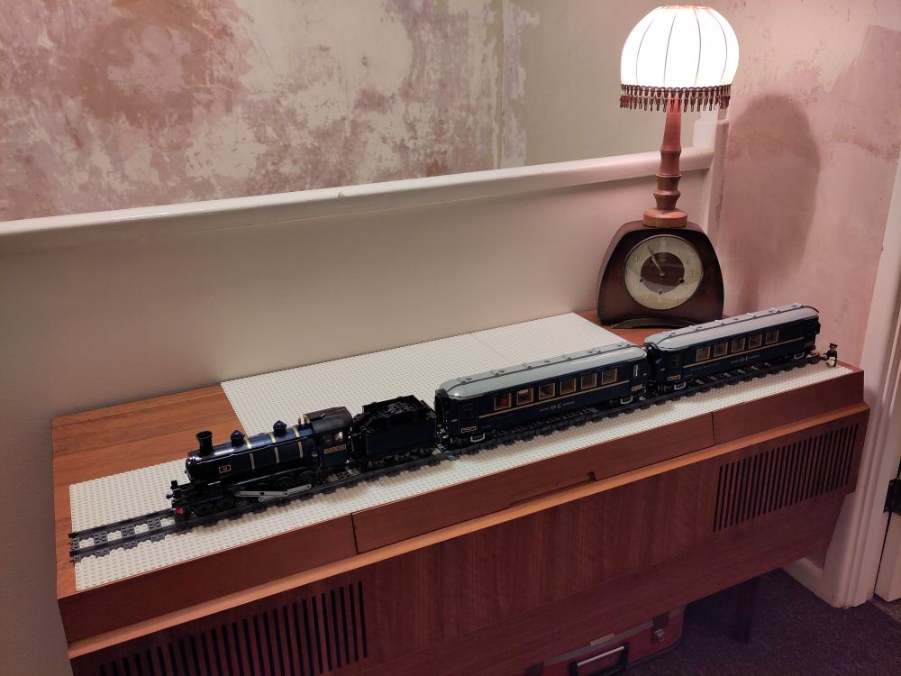 LEGO set 21344 The Orient Express Train stands on a length of railway track upon white baseplates, all on top of a mid-century radiogram. A vintage mantel clock and turned wooden desk lamp stand behind the train.