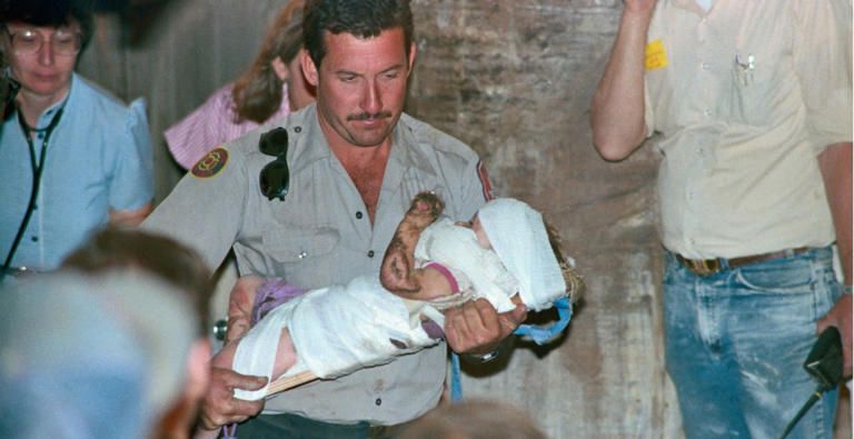 A sheriff or ranger carrying a small child who is strapped to a board because she was just lifted out of a deep well in Texas, 1987