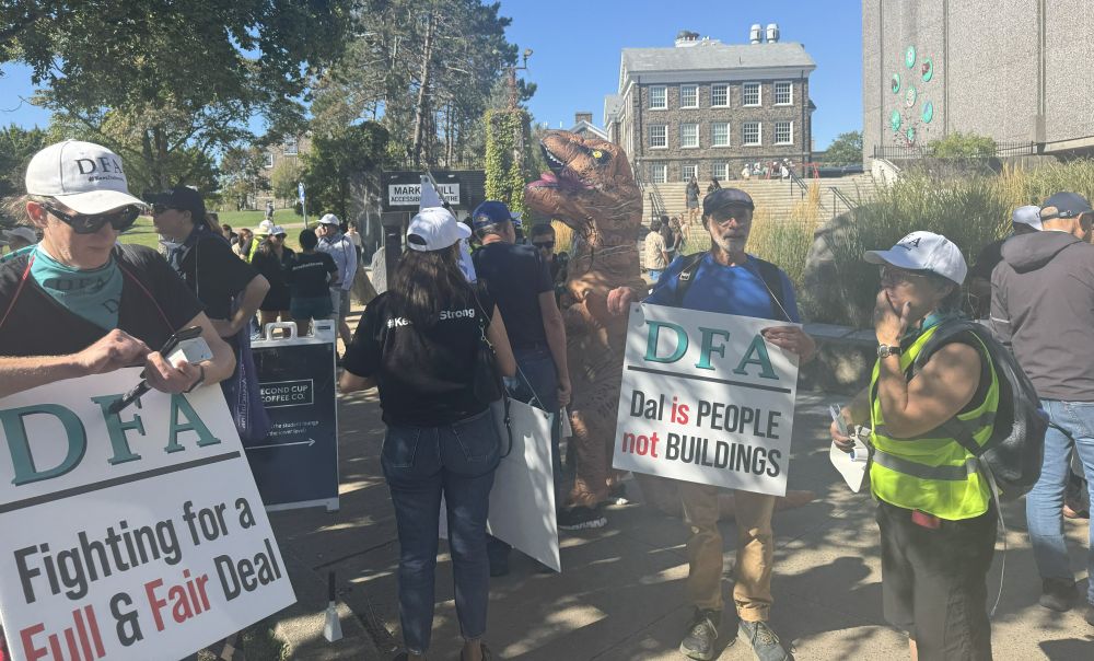Photo of WLUFA's CAUT-DF Representative, Cameron McKenzie posing with a person in an inflatable dinosaur costume on the DFA picket line. He is holding a WLUFA flag