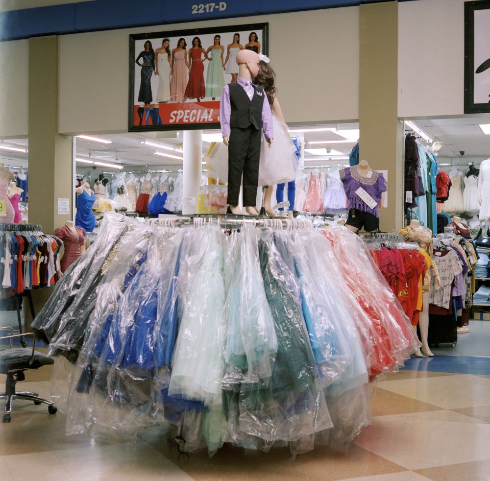 Photograph. A round clothing fixture stuffed with dresses. Two child-sized mannequins stand atop, dressed formal. A clothing shop is visible in the retail space, behind.