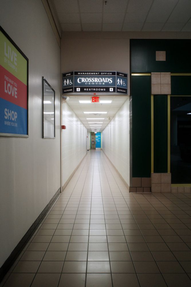 Photograph. Long, empty hallway with a half-open, blue double door at the end. Signage above directs toward the Crossroads Center management offices and restrooms.
