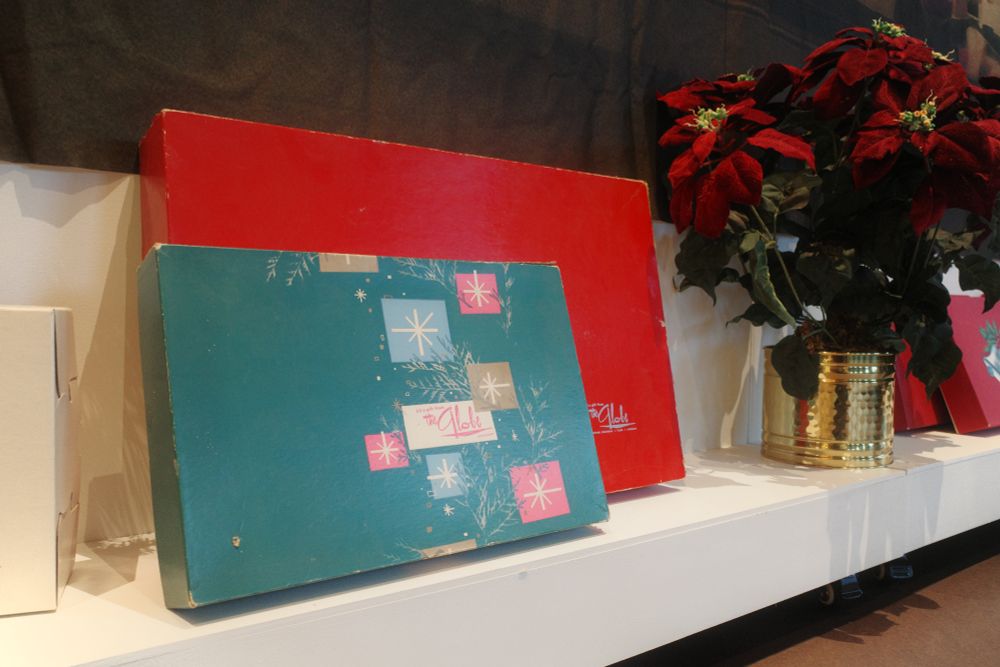 Photograph. Two mid-century gift boxes from the Globe Department Store are on display. Potted poinsetta flowers, to the right. 