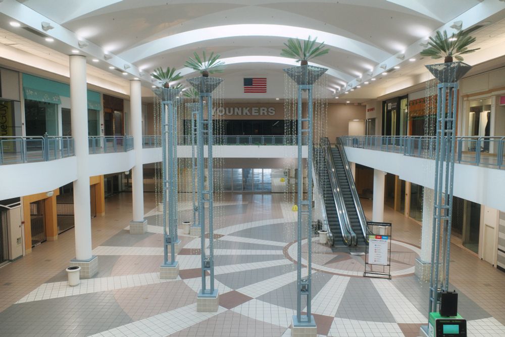 Photograph. Wide-open atrium in an empty, bi-level enclosed mall adorned with turn-of-the-90s decor. Younkers department store anchors the upper level, at the end of the mall.