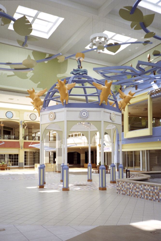 Photograph. A heavily stylized gazebo structure in the central atrium of a dead, enclosed, bilevel shopping mall. There are flying pig statues surrounding the structure and oversized leaf sculptures suspended from the ceiling. The architecture reflects a gaudy Y2K-era renovation completed in 2004.

• Olympus XA4
• Kodak Colorplus 200