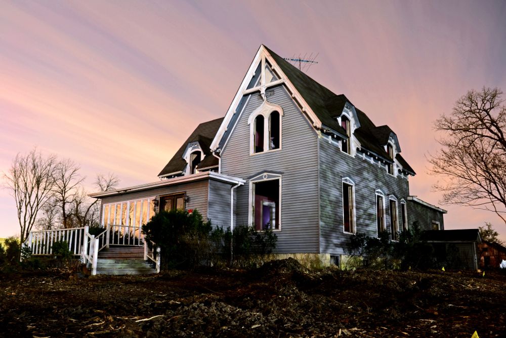 Photograph. A large vacant home surrounded by dead landscape and wood chips from trees that were recently cut down. The home is a two-story farmhouse with an added sunroom and porch on the front. All architectural windows have been removed. Clouds streak the sky in this 17-minute long exposure.