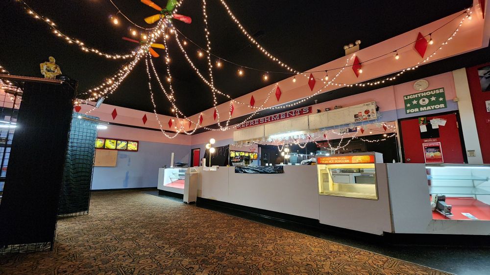 Photograph. A mid-century theater lobby typical of General Cinema Corporation design, albeit with a gaudy paint and carpet job. It is a tall space with string lights strung from the surrounding frieze to colorful ceiling fans. A long concession stand spans the length of the space.