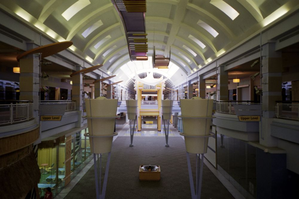 Photograph. A dimly lit concourse in an empty bilevel shopping mall, viewed from the upper level. Oversized decor reflects Y2K-era tastes from a gaudy 2004 renovation.

• Olympus XA4
• Kodak Colorplus 200