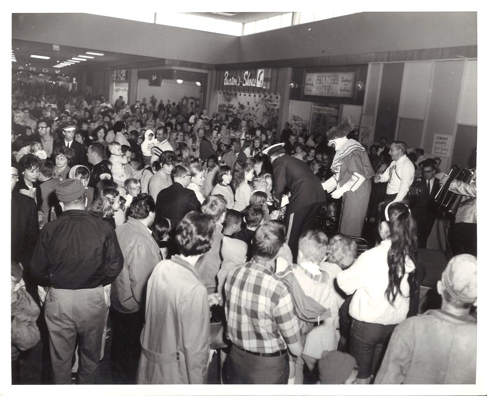 Photograph. An enclosed mall interior lined with stores and filled with people dressed in mid-century fashions, standing shoulder-to-shoulder to meet Bozo the Clown - a television icon in Chicago. Montgomery Wards is visible at the far end of the mall. Other stores visible are Kinney Shoes, Stuart's Fashions, Burton's Shoes, and Printer's Ink. Grand Gardens is under construction behind a temporary wall.
