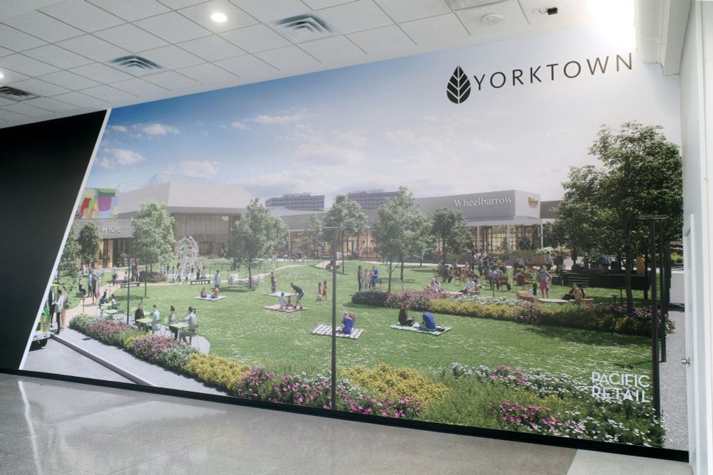 Photograph. A large wall mural depicting concept art for the redevelopment of Yorktown Center mall. It shows a green space with landscaping and the mall in the distance. Fake store names mark the building.