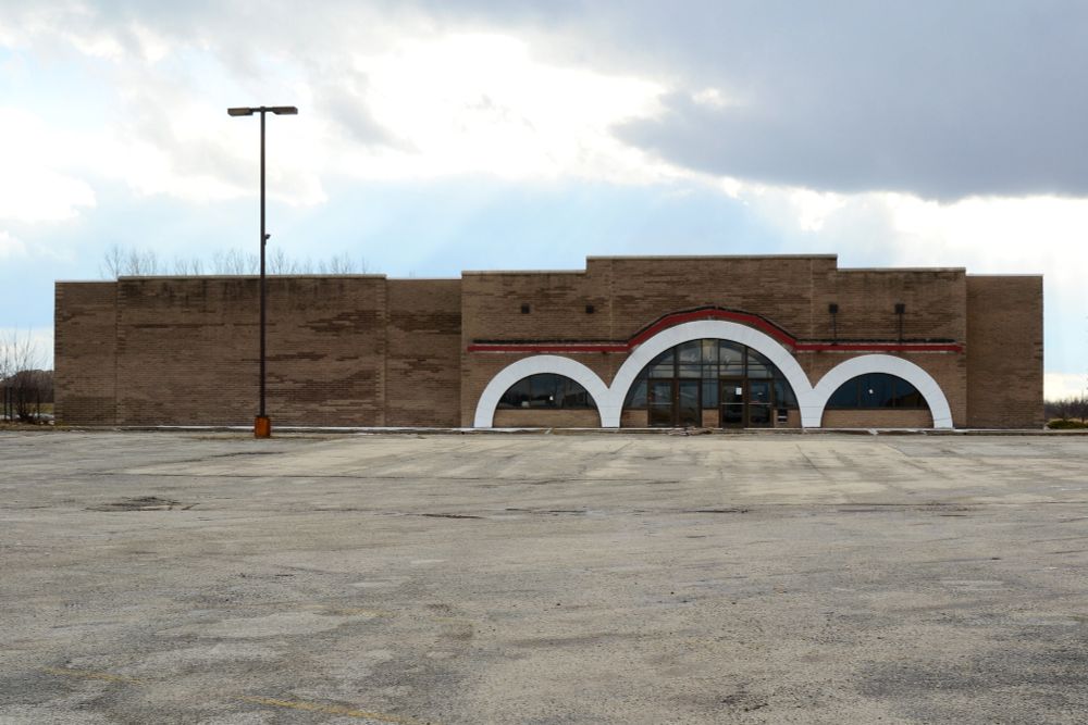 Photograph. A brown brick commercial building originally built for Child World, a defunct toy store chain. There are three white arches surrounding windows with a central doorway serving as the store entrance. The building stands at the end of a crumbling parking lot under overcast skies. A single light pole looms overhead, left of center.