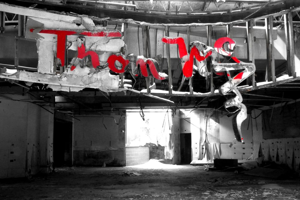 Photograph of a sign for Thom McAn shoes in an abandoned mall. The sign is dangling from a wooden storefront whose structure is sagging from years of water damage, the store itself is gutted.