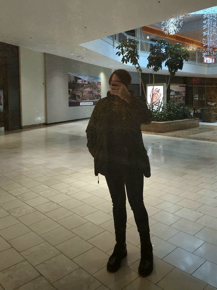 Photograph. Man in black and an army coat. Phone upside down obscuring face. Reflection in a window pane that's been painted from the backside. A dead shopping mall concourse in the background. 