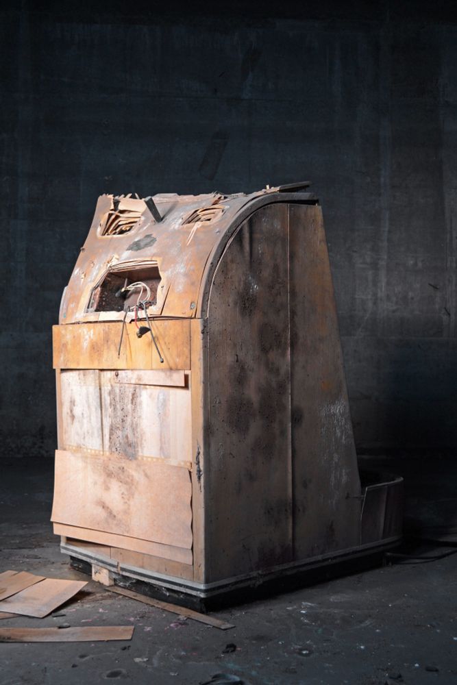 Photograph of a severely distressed and neglected shoe-fitting fluoroscope machine in a dimly-lit concrete room. The device has been stripped of decorative paneling and, x-ray tubes and electronic fittings, leaving openings in the top panel that resemble a screaming face. Debris is scattered across the floor.
