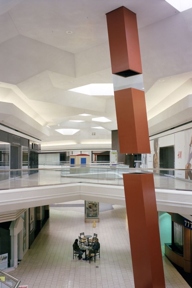Photograph. A towering, slanted red beam with a break in the center, extends floor to ceiling in the concourse of a bilevel shopping center. The mall is dead, no stores are open. Two friends chat at a set of tables on the lower level.