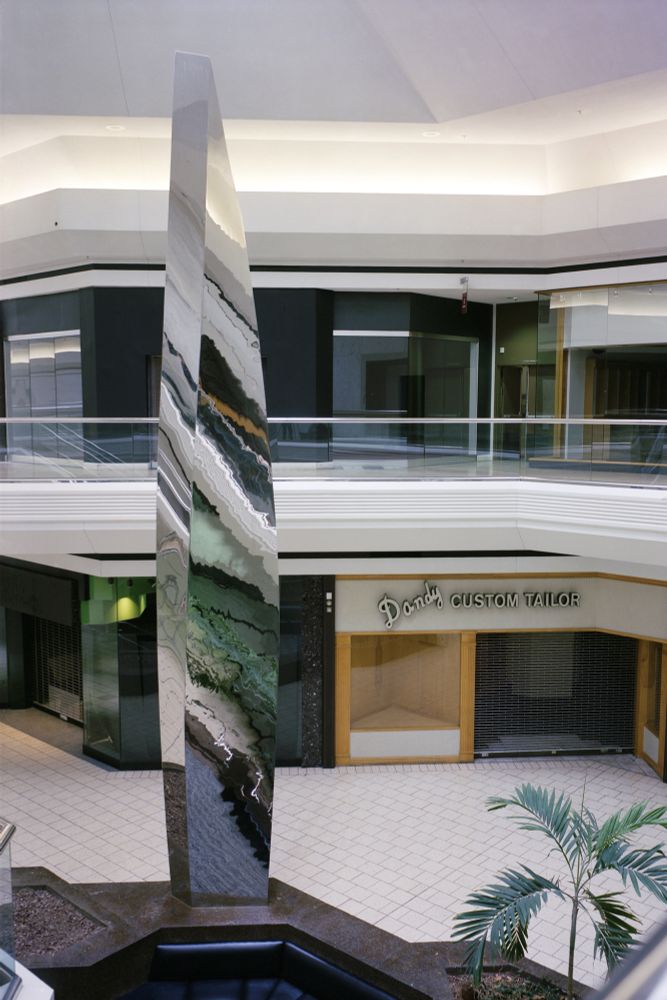 Photograph. A chrome shard of polished metal towers in the atrium of a dead shopping mall. No stores in frame are open, no shoppers are present. The only visible signage on any of the stores is Dandy Custom Tailor on the lower level, right of the sculpture.