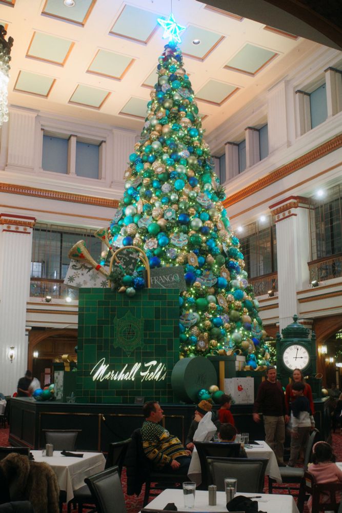 Photograph. A large three-story room housing The Walnut Room restaurant within Macy's flagship department store in Chicago. In 2025 the Great Tree is themed after Marshall Field's, the store which preceded Macy's in this space.