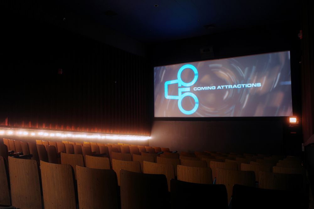 Photograph. An older cinema auditorium with woodgrain-backed seats and string lighting on the far wall. The screen displays the logo of General Cinema Corporation, the theater's original operator during the 1960s through the 1980s, with the words "Coming Attractions"