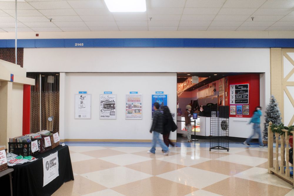 Photograph. A white storefront housing the Belvidere Cinema theater inside Belvidere Mall. Four poster frames hang from the facade, two with historical ads from the theater's opening in 1966; one for the mall's 60th anniversary event; and the last for a free screening of E.T. An entrance is directly right. Mallwalkers pass by the space.