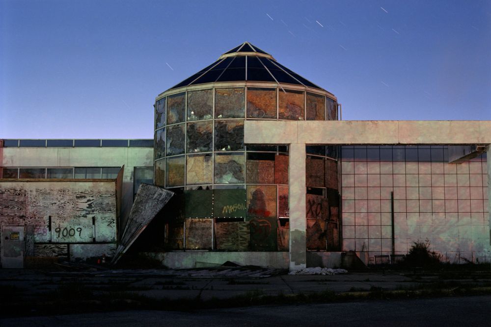 Photograph. An abandoned building at night, a bay window extends from a tile and wood-covered wall, all glass is covered in boards. It is a former entrance to the abandoned Northridge Mall's food court.