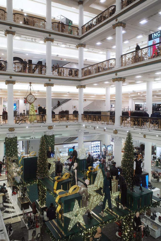 Photograph. Alternate angle of the atrium within Macy's flagship store in Chicago. The aforementioned Holiday display rises over the sales floor. Several floors of merchandise and apparel displays are seen beyond the handrails.