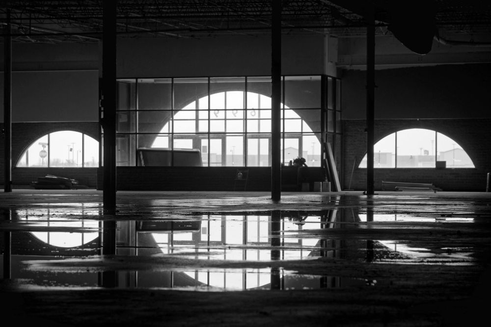 Black and white photograph. Three semi-circular windows allow light into a dark big box retail store. The window shapes were a distinct architectural feature of Child World and Children's Palace, a defunct chain of toy stores whose storefronts were designed to resemble a castle. Water puddles on the concrete floor mirror the image.