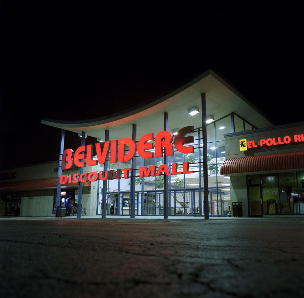 Photograph. A building at night from a very low angle. An inward-curved roof with a glass wall entrance in the center -- signage reads "BELVIDERE DISCOUNT MALL". The centerpiece is flanked by retail space on each side, reflecting more 90s architecture with quarter-circle awnings made of corrugated steel.
