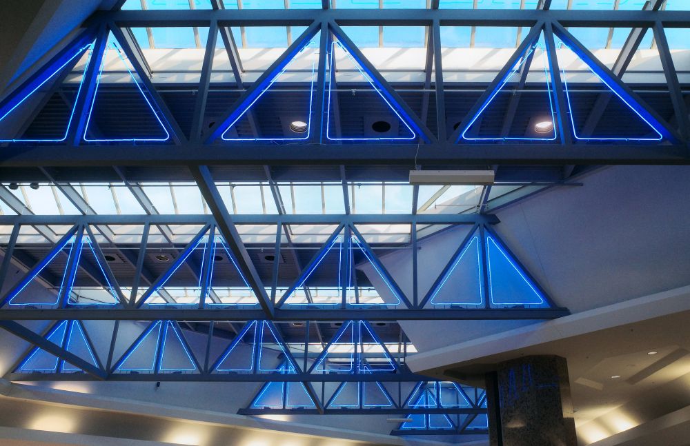 Photograph. Ceiling detail over the center court of a shopping mall. The ceiling features skylights and oversized trusses, which are accented by triangular-shaped blue neon designed tor resemble sailboats on the horizon. Warmer lighting accents the soffits, below.