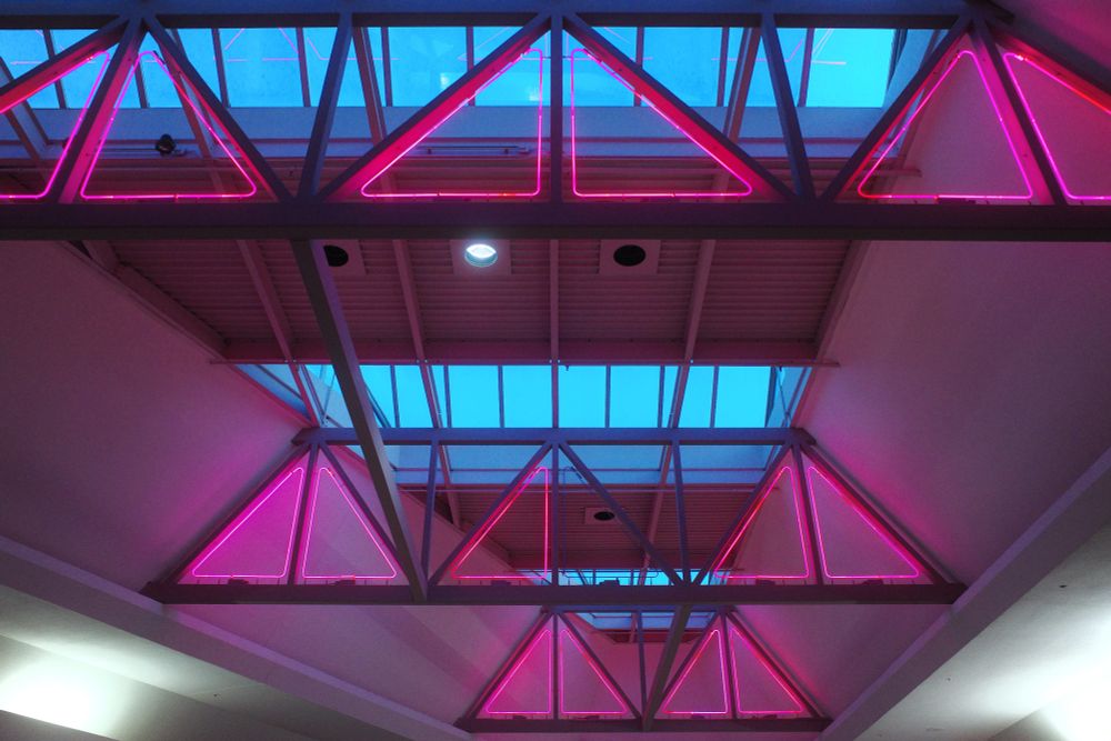 Photograph. Detail view of a skylight overhead a mall atrium. The glass ceiling is broken by sections of metal roofing, and oversized trusses featuring illuminated pink neon triangle inserts.
