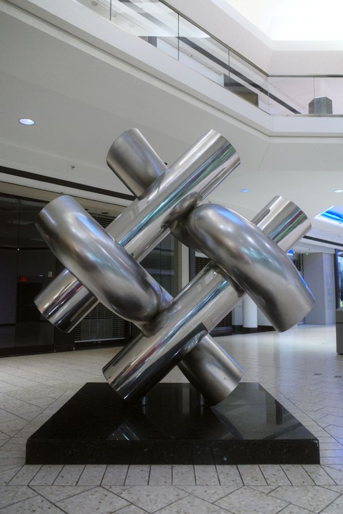 Photograph. A large, polished metal sculpture in a dead, enclosed shopping mall. The sculpture consists of four steel tubes -- two curved tubes wrapping around two straight tubes -- angled at 45-degrees. The sculpture is mounted to a black marble pedestal surrounded by terrazzo tiles. Empty retail storefronts are visible in the background.