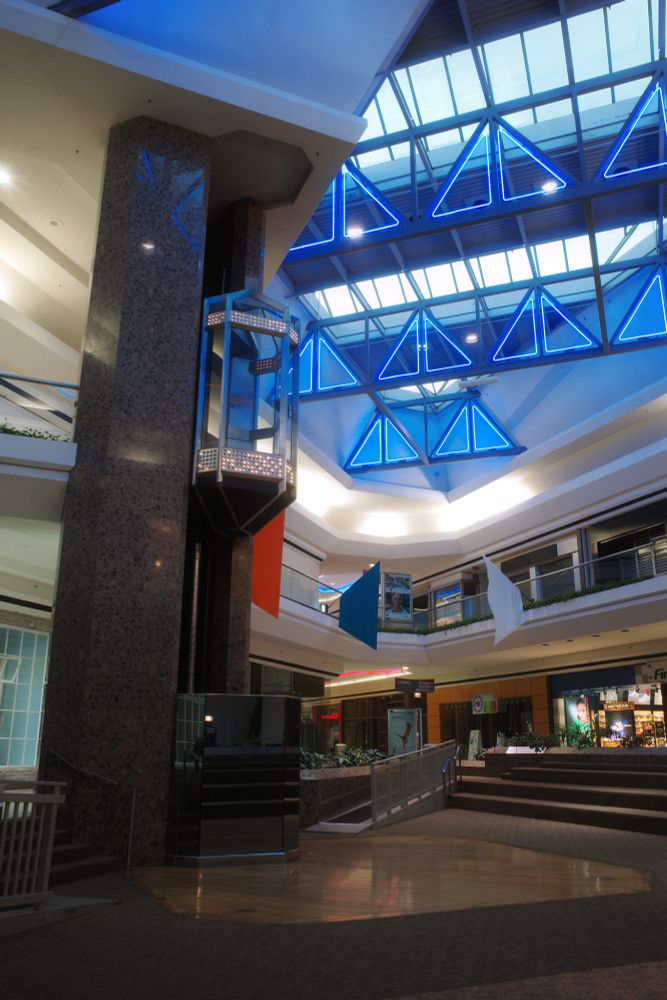 Photograph. A bilevel shopping mall's center court -- it has a postmodern pod elevator with the car on the upper level, and an amphitheater stage at its base. Most of the retail shops on both level are dark except for Finish Line. The ceiling features skylights and oversized trusses, which are accented by triangular-shaped blue neon designed tor resemble sailboats on the horizon. Solid-colored flags hang from the balcony.